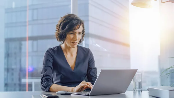 Selbstbewusste Geschäftsfrau, die an ihrem Schreibtisch sitzt und in ihrem modernen Büro an einem Laptop arbeitet. Stilvolle schöne Frau, die einen wichtigen Job macht. Im Fenster Großstadt-Business-District-Blick mit Sonnenstrahl. — Stockfoto