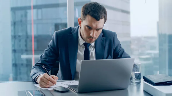 Успешный бизнесмен сидит за своим столом, работает с документами, жестко подписывает документы и смотрит на ноутбук. Вид из окна на деловой район большого города. — стоковое фото