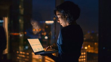 Başarılı, şık giyimli iş kadını ofisinin penceresinin yanında dikilirken dizüstü bilgisayar tutuyor. Gecenin geç saatlerinde Profesyonel Kadın Önemli İş Yapıyor 'da. Pencere Büyük Şehir İş Bölgesi Görünümüne sahip