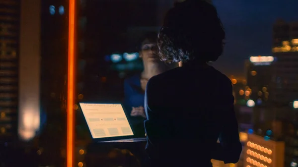 Elegantemente vestida mujer de negocios sostiene el ordenador portátil mientras mira por la ventana de su oficina. Tarde en la noche Mujer haciendo un trabajo importante. Ventana tiene vista del distrito de negocios de la ciudad grande con muchas luces nocturnas. —  Fotos de Stock