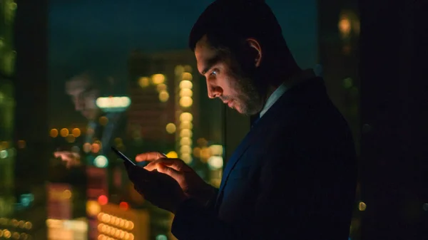 Tarde en la noche en la oficina Negocios exitosos Tipos de correo electrónico en el teléfono inteligente. En la ventana Vista del distrito de negocios con luces de la ciudad. —  Fotos de Stock