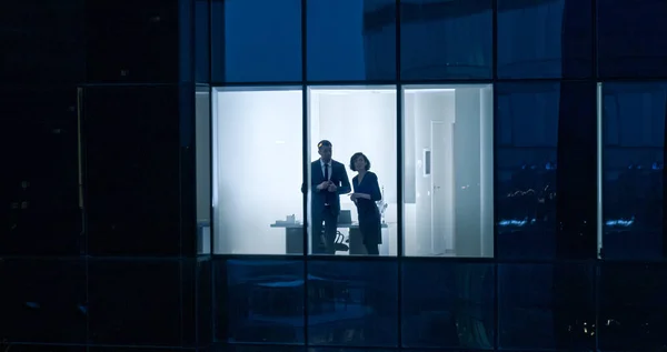 Aerial Shot From Outside of the Skyscraper: Businessman and Businesswoman Talking while Looking Out of the Office Window (en inglés). Disparo del Distrito Comercial en la Noche y Empresarios Trabajando en el —  Fotos de Stock