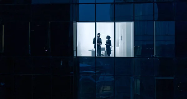Aerial Shot From Outside of the Skyscraper: Businessman and Businesswoman Talking while Standing in the Office Window (em inglês). Tiro do distrito de negócios na noite e empresários que trabalham no — Fotografia de Stock