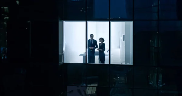 Flygfotografering utanför skyskrapan: Affärsman och affärskvinna som pratar medan de tittar ut genom kontorsfönstret. Skjuten av affärsdistriktet i kväll och affärsmän som arbetar i — Stockfoto