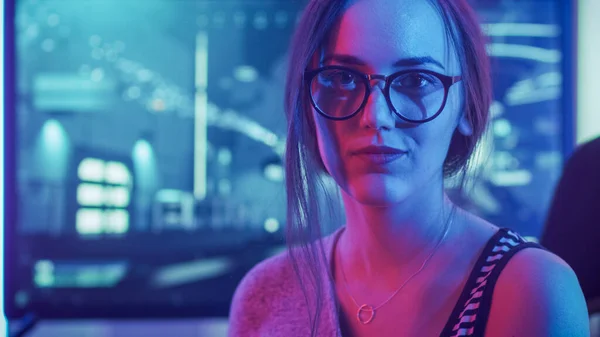 Portrait of the Beautiful Young Pro Gamer Girl Sitting at Her Personal Computer and Looks into Camera Smiling. Attractive Geek Girl Player Wearing Glasses in the Room Lit by Neon Lights.