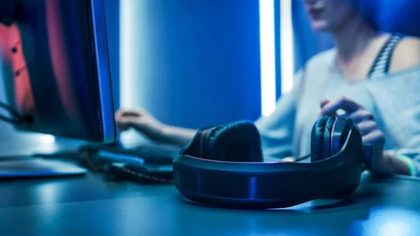 Close-up Shot of a Pro Gaming Headset. In the Background Beautiful Professional Gamer Girl Starts Playing Online Video Game on Her Personal Computer. Cute Casual Geek Girl. Room Lit by Neon Lamps in — Stock Photo, Image