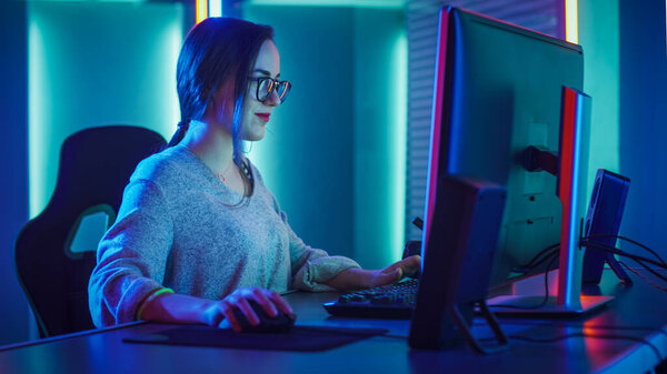 Beautiful Professional Gamer Girl Sitting Down to Play in First-Person Shooter Online Video Game on Her Personal Computer. Casual Cute Geek wearing Glasses and Talking into Headset. Cyber e-Sport