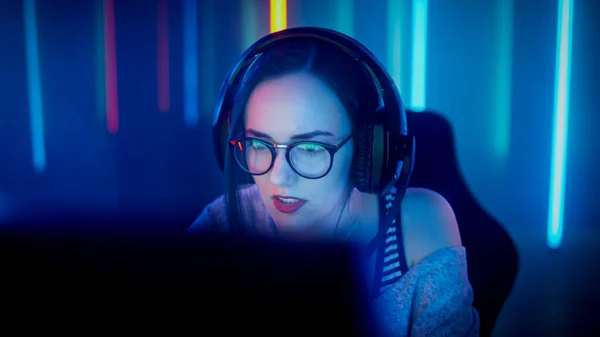 美丽的职业游戏女孩在她的个人电脑上玩网络游戏。带着眼镜和对着麦克风说话的随意可爱的极客。网上体育比赛. — 图库照片
