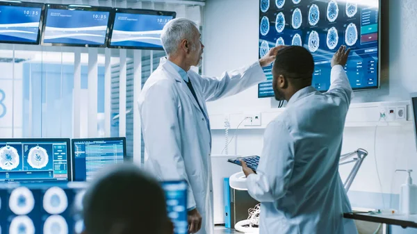 Beyin Araştırma Laboratuvarı 'nda Bilim Adamları Ekibi, Duvar TV Monitöründe Beyin Taramaları Gösterisini tartışıyor. Nörologlar etrafı monitörlerle çevrili, tomografi ve MRI taramaları gösteriyor..