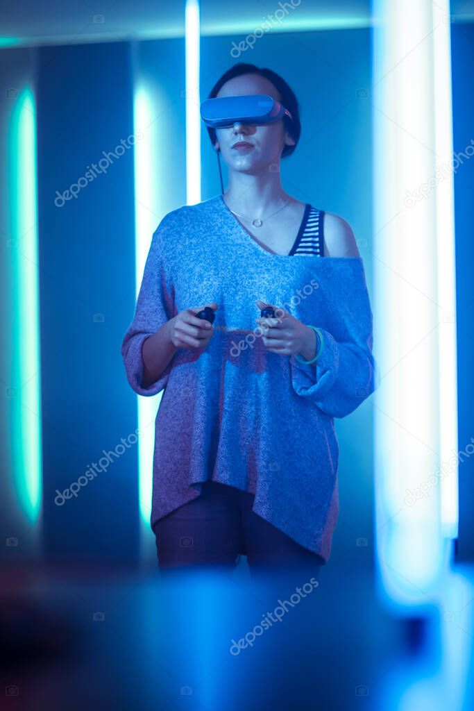 Beautiful Young Girl Wearing Virtual Reality Headset Ready to Play and Holding Joysticks Controllers. Creative Young Girl Playing Online Video Game. Neon Retro Lights Surround Her. Vertical Photo.