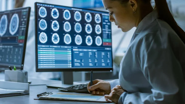 Kvinnlig medicinsk forskare Arbetar med hjärnskannrar på hennes persondator, skriva ner data i en Urklipp. Modernt laboratorium som arbetar med neurofysiologi, vetenskap, neurofarmakologi. — Stockfoto