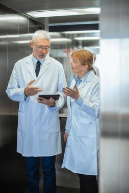 Hastanedeki iki doktor asansörde duruyor ve Tablet Bilgisayar kullanırken tartışıyorlar. Yeni Modern Tamamen İşlevsel Tıbbi Tesis.