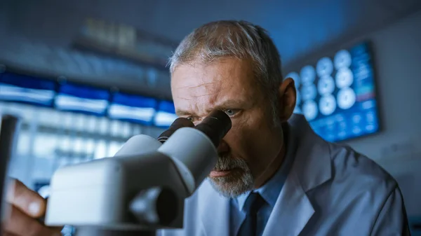 Senior Medical Research Scientist Looking under the Microscope in the Laboratory. Neurologist Solving Puzzles of the Mind and Brain. In the Laboratory with Multiple Screens Showing MRI CT Brain Scan