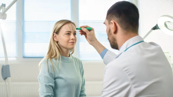 Sobre el hombro de un cirujano cosmético plástico dibuja líneas en una cara de un hermoso modelo femenino para la operación de lifting facial. —  Fotos de Stock