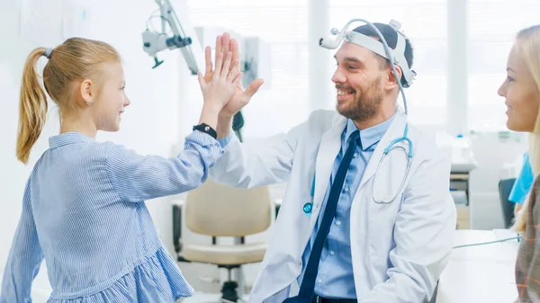 Pediatra de ORL amigable le da cinco a una niña sonriente después del examen de salud con el uso de un equipo especial de linterna. Madre con una pequeña hija Visite el consultorio de médicos. —  Fotos de Stock