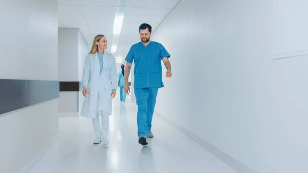 Хирург и женщина-доктор в спешке проходят по больничному коридору, говоря о здоровье пациентов. Современная светлая больница с профессиональным персоналом. — стоковое фото