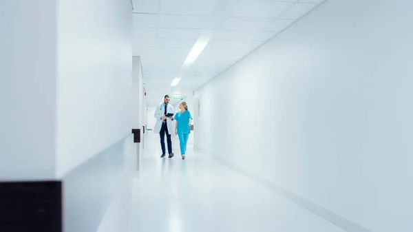Хирург и женщина-врач в спешке проходят по больничному коридору, используя цифровые таблетки и говоря о здоровье пациентов. Современная светлая больница с профессиональным персоналом. — стоковое фото