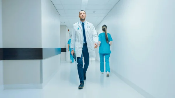 Beslutsam stilig doktor bär vit rock med stetoskop går genom sjukhuset Hallway. Modern ljus klinik med professionell personal. — Stockfoto