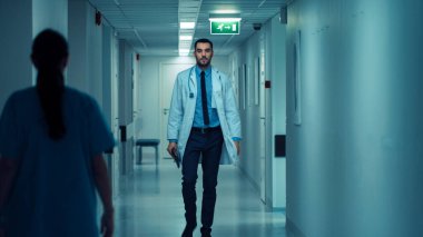Hastane koridorunda steteskopla yürüyen kararlı, yakışıklı doktor, beyaz önlüklü. Profesyonel Personelle Modern Parlak Klinik.