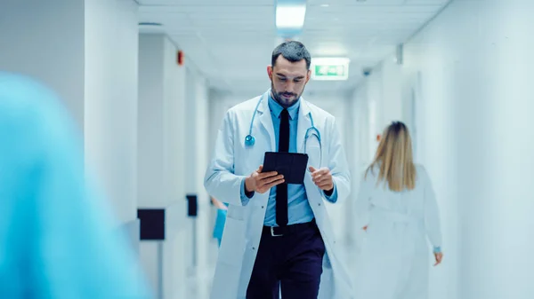 Déterminé beau médecin utilise ordinateur tablette numérique tout en marchant dans le couloir de l'hôpital. Clinique lumineuse moderne avec personnel professionnel. — Photo