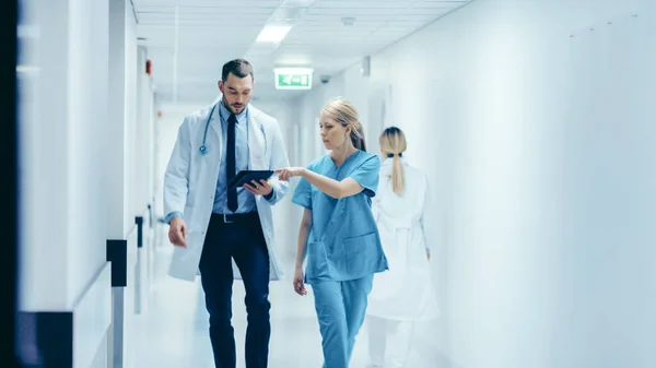 여성 의사와 의사는 병원 복도를 걸으며, 환자 건강에 대해 이야기하는 동안 디지털 태블릿 컴퓨터를 상담 한다. 프로페셔널 스태프들과 함께 한 현대 브라이트 병원. — 스톡 사진