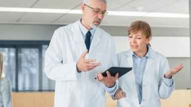 Hastane doktorları lobide yürürken, Tablet Bilgisayar kullanırken tartışın. Geçmiş Hastalar ve Tıbbi Personel 'de. Yeni Modern Tamamen İşlevsel Tıbbi Tesis.