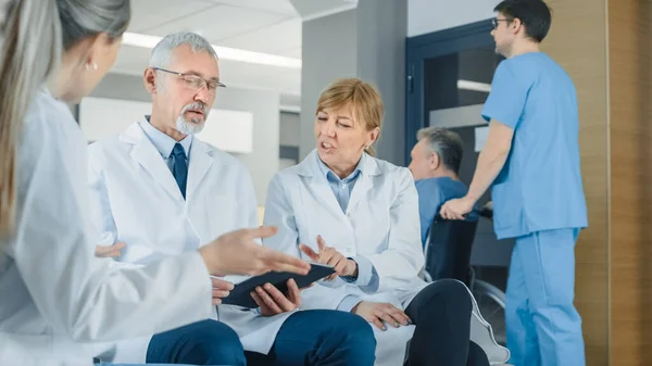 병원의 로비에서 세 명의 전문 의사가 앉아 있는 동안 토론을 한다. 그들은 태블릿 컴퓨터를 사용 한다. 시골에 최고의 전문 의료인들과 의료 원이 있는 바쁜 현대 병원. — 스톡 사진