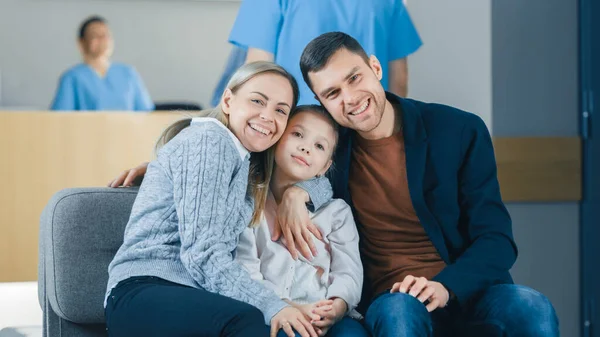 Portrait of the Young Happy Family in the Hospital. Handsome Father, Beautiful Mother and Cute Little Daughter Sitting in the Lobby of the Medical Facility, Smiling. — Stock Photo, Image