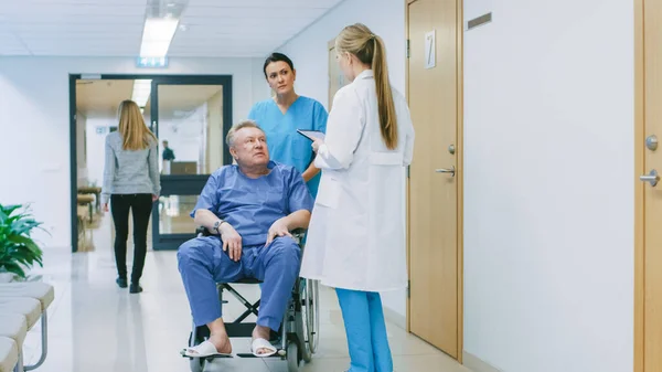En el pasillo del hospital, la enfermera empuja al paciente anciano en silla de ruedas, el doctor les habla mientras usa la computadora de la tableta. Limpio, nuevo hospital con personal médico profesional. —  Fotos de Stock