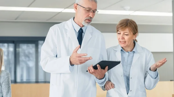 Im Krankenhaus gehen Ärzte durch die Lobby, diskutieren, während sie Tablet-Computer benutzen. Im Hintergrund Patienten und medizinisches Personal. Neue moderne, voll funktionsfähige medizinische Einrichtung. — Stockfoto