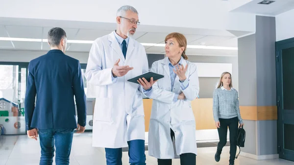 No hospital, médicos ocupados conversam, usando o Tablet Computer enquanto caminham pelo prédio. No fundo, o paciente conversa com o recepcionista. Nova instalação médica moderna totalmente funcional. — Fotografia de Stock