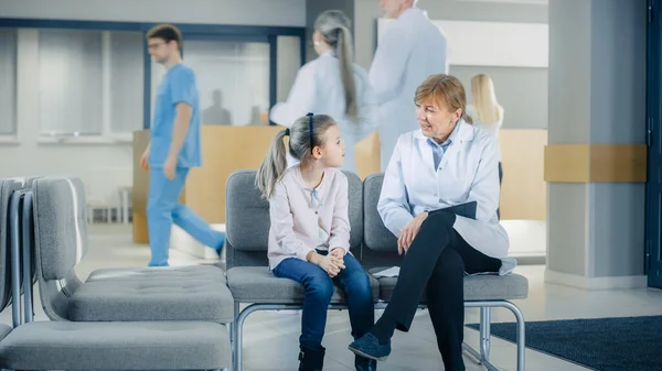 In de lobby van het ziekenhuis praat een vrouwelijke dokter met een schattig klein meisje terwijl ze op de bank zit. Drukke moderne ziekenhuis met best mogelijke kindergeneeskunde afdeling in het land. — Stockfoto