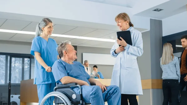 病院のロビーでは、車椅子の高齢者を押している看護師、医師がタブレットコンピュータを使用している間に彼らに話します。専門医療従事者とのクリーン、新しい病院. — ストック写真