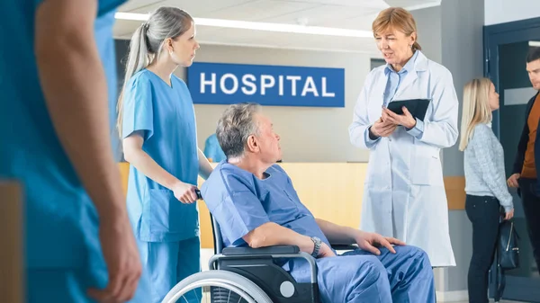 En el vestíbulo del hospital, la enfermera empuja al paciente anciano en silla de ruedas, el doctor les habla mientras usa la computadora de la tableta. Limpio, nuevo hospital con personal médico profesional. —  Fotos de Stock