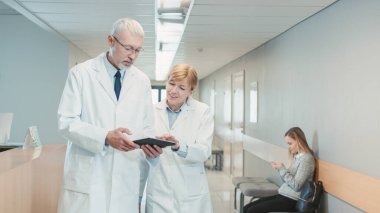 Hastane koridorunda yürüyen iki doktor konuşuyor ve tablet bilgisayarı paylaşıyor. Hastalar ve Tıbbi Personelle Modern Hastane.