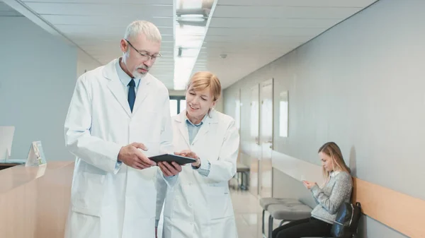 W szpitalu dwaj lekarze spacerujący korytarzem szpitalnym rozmawiają i dzielą się tabletem. Nowoczesny szpital z pacjentami i personelem medycznym. — Zdjęcie stockowe