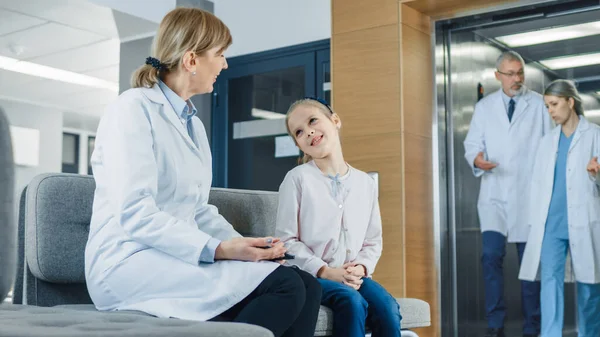 In de lobby van het ziekenhuis praat een vrouwelijke dokter met een schattig klein meisje terwijl ze op de bank zit. Drukke moderne ziekenhuis met best mogelijke kindergeneeskunde afdeling in het land. — Stockfoto