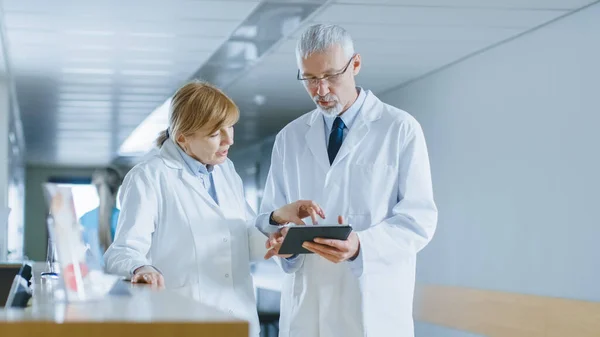 I sjukhuset två läkare står vid receptionen, prata och dela information på Tablet Computer. Modernt sjukhus med patienter och medicinsk personal. — Stockfoto