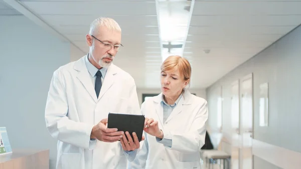 Im Krankenhaus gehen zwei Ärzte durch den Krankenhausflur, unterhalten sich und teilen Tablet-Computer. Modernes Krankenhaus mit Patienten und medizinischem Personal. — Stockfoto