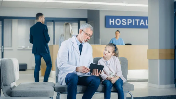 In de lobby van het ziekenhuis spreekt een mannelijke arts met een schattig klein meisje terwijl ze op de bank zit, hij toont haar tablet computer. Drukke moderne ziekenhuis met best mogelijke kindergeneeskunde afdeling in — Stockfoto