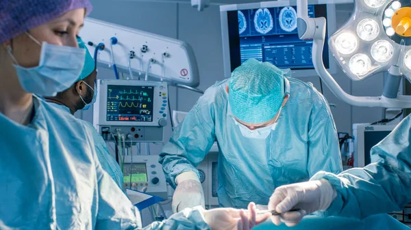 Diverse Team of Professional Surgeons Performing Invasive Surgery on a Patient in the Hospital Operating Room. Nurse Hands Out Instruments to surgeon, Anesthesiologist Monitors Vitals. Real Modern