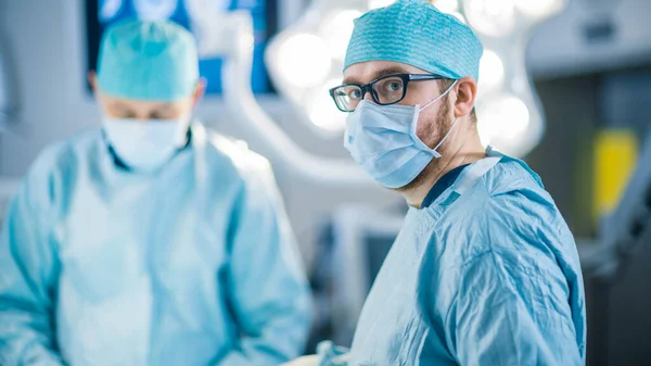 Porträtaufnahme eines Chirurgen, der in die Kamera blickt. Vielfältiges Team professioneller Chirurgen, Assistenten und Krankenschwestern, die invasive Operationen an einem Patienten im Operationssaal des Krankenhauses durchführen. — Stockfoto