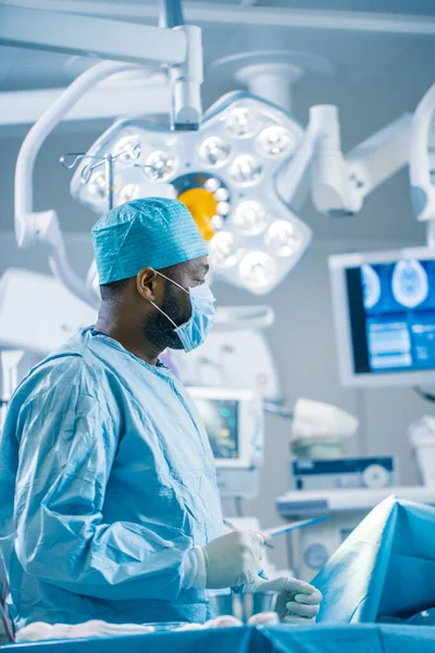 Portrait of the Black Professional Surgeon Performing Invasive Surgery on a Patient in the Hospital Operating Room. In the Background Modern Hospital Operating Room. Vertical Shot.