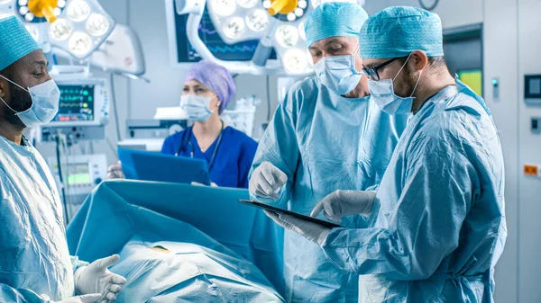 Cirujanos y asistentes profesionales hablan y usan Tablet Computer digital durante la cirugía. Trabajan en la sala de operaciones del hospital moderno. —  Fotos de Stock