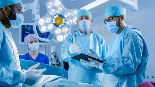 Professionelle Chirurgen und Assistenten sprechen und verwenden während der Operation digitale Tablet-Computer. Sie arbeiten im modernen Operationssaal des Krankenhauses. — Stockfoto