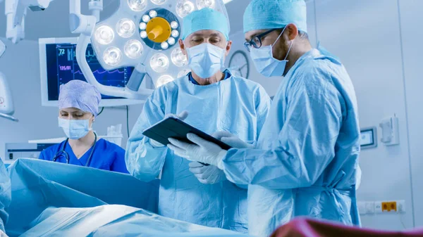 Professionelle Chirurgen und Assistenten sprechen und verwenden während der Operation digitale Tablet-Computer. Sie arbeiten im modernen Operationssaal des Krankenhauses. — Stockfoto