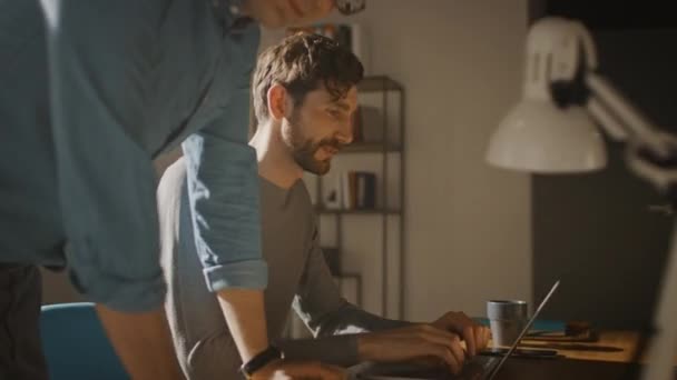 Profesional Creative Man Sitting at His Desk works on a Laptop, His Colleague Helps with Poignant Advice, They Have Discussion about Decisions made for Project (dalam bahasa Inggris). 360 Degree Tracking Arc Shot — Stok Video