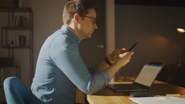 Professionell Creative Man Sitter vid sitt skrivbord i Home Office Studio Arbetar på en bärbar dator. Man Använda Smartphone för att kontrollera sociala medier, Sök Internet. 360 graders spårning båge skott rörelse — Stockvideo