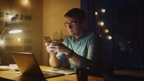 Profesjonalista siedzi przy biurku w studiu biurowym Wieczorem pracuje na laptopie. Człowiek korzystający ze smartfona z danymi, analizujący statystyki. 360 Stopień śledzenia ruchu łuku strzał — Wideo stockowe