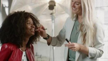 Fotoğraf çekiminin sahne arkası: Makyöz, gür kıvırcık saçlı güzel siyah modele ruj sürüyor. Moda Dergisi Stüdyosu Fotoğraf Hazırlıkları. Profesyonel Ekipman Stüdyosu